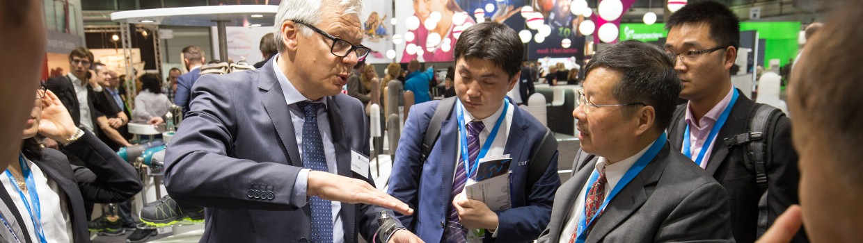 Ein Mann im Anzug erklärt einer Gruppe von Messebesuchern ein technisches Produkt an einem Ausstellungsstand. Ein Mann im Anzug erklärt einer Gruppe von Messebesuchern ein technisches Produkt an einem Ausstellungsstand.