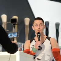 Ein Mann im Sporttrikot mit roten Kompressionsärmeln spricht in ein Mikrofon; im Vordergrund filmen zwei Personen mit Smartphones