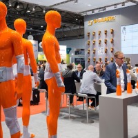 Messestand von SPORLASTIC auf der OTWorld mit zahlreichen orangefarbenen Ganzkörper-Dummies, die Orthesen an Armen, Beinen und Rumpf tragen; im Vordergrund sprechen zwei Personen an einem Stehtisch, im Hintergrund sitzen weitere Personen an Tischen, ein großer Bildschirm zeigt ein Produktbild.