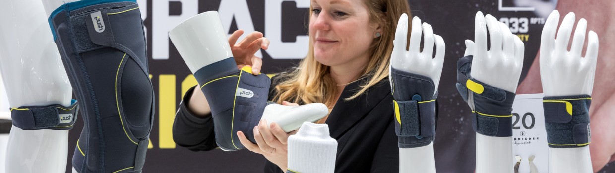 Eine Frau demonstriert am Messestand auf der OTWorld eine Sprunggelenkorthese an einem weißen Modellbein mit gelbem Sportschuh; auf dem Tisch stehen weitere Modellarme und -beine mit grauen Orthesen der Marke PUSH, im Hintergrund ist der Schriftzug „ANKLE BRACE“ sichtbar. Eine Frau demonstriert am Messestand auf der OTWorld eine Sprunggelenkorthese an einem weißen Modellbein mit gelbem Sportschuh; auf dem Tisch stehen weitere Modellarme und -beine mit grauen Orthesen der Marke PUSH, im Hintergrund ist der Schriftzug „ANKLE BRACE“ sichtbar.