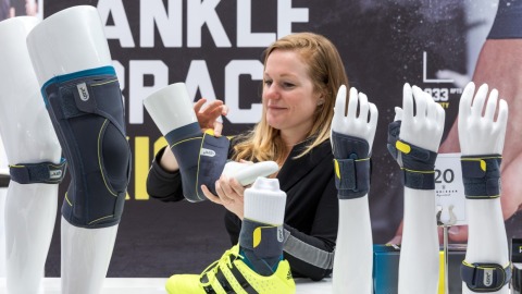 Eine Frau demonstriert am Messestand auf der OTWorld eine Sprunggelenkorthese an einem weißen Modellbein mit gelbem Sportschuh; auf dem Tisch stehen weitere Modellarme und -beine mit grauen Orthesen der Marke PUSH, im Hintergrund ist der Schriftzug „ANKLE BRACE“ sichtbar.