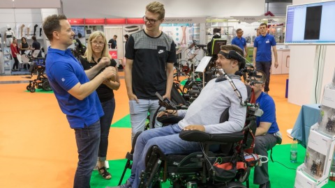 A woman demonstrates an ankle brace on a white model leg with a yellow sports shoe at the OTWorld booth; on the table are additional model arms and legs fitted with gray braces by the brand PUSH, with the text “ANKLE BRACE” visible in the background.