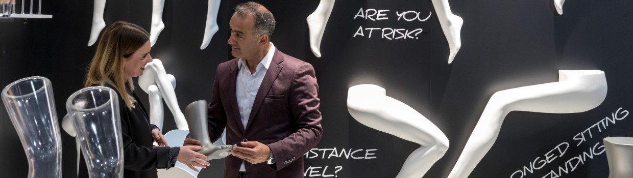 Zwei Personen stehen auf der OTWorld vor einer schwarzen Ausstellungswand mit weißen Modellbeinen und großem Text wie „VARICOSE VEINS?“ und „PROLONGED SITTING OR STANDING?“; sie betrachten gemeinsam ein Ausstellungsstück in Beinform. Zwei Personen stehen auf der OTWorld vor einer schwarzen Ausstellungswand mit weißen Modellbeinen und großem Text wie „VARICOSE VEINS?“ und „PROLONGED SITTING OR STANDING?“; sie betrachten gemeinsam ein Ausstellungsstück in Beinform.
