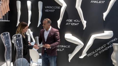 Zwei Personen stehen auf der OTWorld vor einer schwarzen Ausstellungswand mit weißen Modellbeinen und großem Text wie „VARICOSE VEINS?“ und „PROLONGED SITTING OR STANDING?“; sie betrachten gemeinsam ein Ausstellungsstück in Beinform.