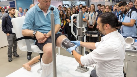 Ein Mann sitzt auf der OTWorld auf einem Stuhl, während ihm ein Orthopädietechniker einen durchsichtigen Guss für eine Beinprothese anpasst; zahlreiche Messebesucher beobachten die Demonstration im Hintergrund.