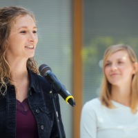 Eine Frau spricht ins Mikro. Eine weitere weibliche Person steht im Hintergrund.