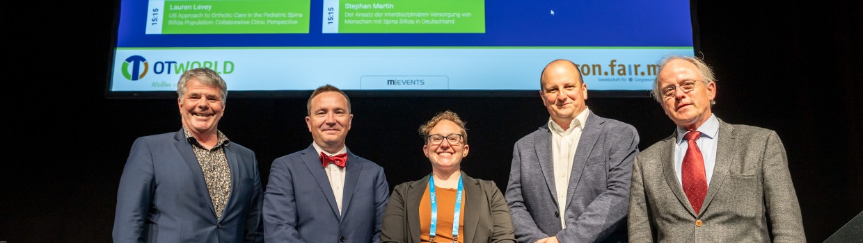 Fünf Personen stehen nebeneinander auf einer Bühne und blicken in die Kamera. Im Hintergrund ist eine große Leinwand mit Präsentationsfolien zur orthopädischen Versorgung zu sehen. Die Personen tragen Konferenz-Badges. Fünf Personen stehen nebeneinander auf einer Bühne und blicken in die Kamera. Im Hintergrund ist eine große Leinwand mit Präsentationsfolien zur orthopädischen Versorgung zu sehen. Die Personen tragen Konferenz-Badges.