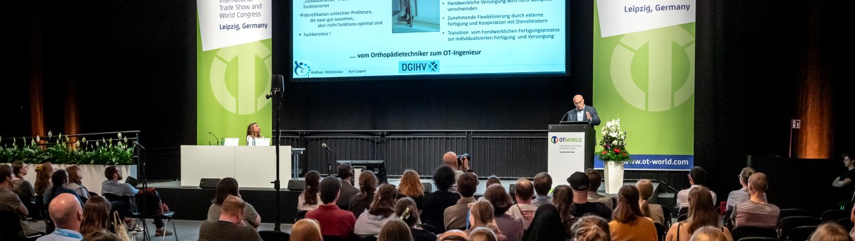 Eine Vortragsveranstaltung bei der OTWorld in Leipzig. Zahlreiche Teilnehmende sitzen im Publikum und verfolgen die Präsentation eines Sprechers auf der Bühne. Auf einer großen Leinwand ist eine Folie mit Informationen sichtbar, flankiert von zwei OTWorld-Bannern.