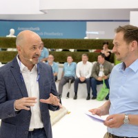 Two men in conversation at OtWorld, one has a microphone, the other is holding a pad with notes. In the background, several people are sitting and standing in front of an exhibition area. Two men in conversation at OtWorld, one has a microphone, the other is holding a pad with notes. In the background, several people are sitting and standing in front of an exhibition area.