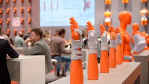 Several orange arm prostheses with gray orthoses are displayed in a row on a table; in the background, people are seated at tables, with more orange torso dummies on the wall and a large screen showing a presentation.