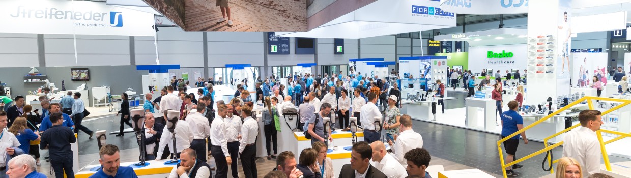 View of a trade fair hall with numerous visitors and crowded booths of various companies, including “DARCO,” “Streifeneder,” and “Basko”; large advertising banners showing people with prostheses in everyday settings hang from the ceiling.