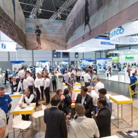 View of a trade fair hall with numerous visitors and crowded booths of various companies, including “DARCO,” “Streifeneder,” and “Basko”; large advertising banners showing people with prostheses in everyday settings hang from the ceiling. View of a trade fair hall with numerous visitors and crowded booths of various companies, including “DARCO,” “Streifeneder,” and “Basko”; large advertising banners showing people with prostheses in everyday settings hang from the ceiling.