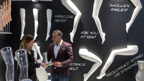 Two people stand at OTWorld in front of a black display wall with white model legs and large text such as “VARICOSE VEINS?” and “PROLONGED SITTING OR STANDING?”; they are examining a leg-shaped exhibit together.