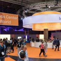 Visitors walk through the Congress Center at OTWorld, featuring an orange floor and a large circular information booth above which hangs signage for congress bags and tickets; to the left, a large banner reads “Congress Center OTWorld.” Visitors walk through the Congress Center at OTWorld, featuring an orange floor and a large circular information booth above which hangs signage for congress bags and tickets; to the left, a large banner reads “Congress Center OTWorld.”