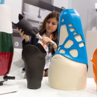 Several colorful and partially open-structured leg prosthetic sockets are displayed on a table at OTWorld; in the background, a woman is holding another black prosthetic model. Several colorful and partially open-structured leg prosthetic sockets are displayed on a table at OTWorld; in the background, a woman is holding another black prosthetic model.
