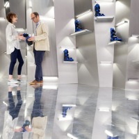 Two people stand in a modern exhibition space at OTWorld with a reflective floor and illuminated wall niches displaying blue orthoses; both are holding an orthosis and engaged in conversation. Two people stand in a modern exhibition space at OTWorld with a reflective floor and illuminated wall niches displaying blue orthoses; both are holding an orthosis and engaged in conversation.