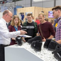 An exhibitor at OTWorld explains various orthopedic therapy shoes to a group of young people, with the shoes displayed on white decorative stones; in the background, other visitors and a mannequin wearing a back brace are visible. An exhibitor at OTWorld explains various orthopedic therapy shoes to a group of young people, with the shoes displayed on white decorative stones; in the background, other visitors and a mannequin wearing a back brace are visible.