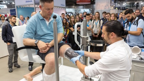 A man sits on a chair at OTWorld while an orthopedic technician fits a transparent mold for a leg prosthesis; numerous trade fair visitors are observing the demonstration in the background.