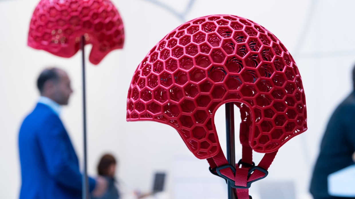 Red protective helmet with honeycomb structure in the foreground, displayed at OTWorld, with trade fair visitors in the background. Red protective helmet with honeycomb structure in the foreground, displayed at OTWorld, with trade fair visitors in the background.