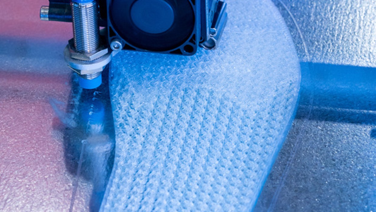A 3D printer is printing an orthopedic insole made of light-colored material on a metal platform; visible are the print head with cables and the forming object under blue lighting. A 3D printer is printing an orthopedic insole made of light-colored material on a metal platform; visible are the print head with cables and the forming object under blue lighting.
