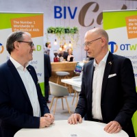 Two men are talking at a bar table. The OTWorld logo can be seen on adisplay in the background.