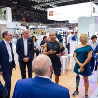 Group of people gathered at the OT-World trade fair around two athletic individuals with prostheses, engaged in lively discussion within a modern exhibition area.