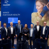 Group photo of trade fair visitors at the Thuasne booth at OTWorld, in front of a blue wall with the slogan ‘Wings for your health’ and a large image of a woman wearing a wrist brace.