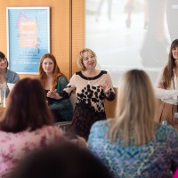 Four women on a panel speaking during a presentation, one woman standing and actively moderating in front of an audience.