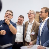 Group of professionals at OTWorld enthusiastically watching a product demonstration, several people smiling and engaged in conversation.