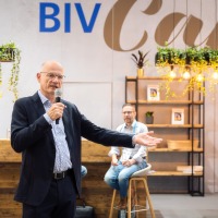 Man speaking into a microphone at the BIV Café during OTWorld, with a cozy café setting in the background including bar stools, plants, and shelves