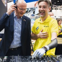 Man with prosthetic arm explains DJ equipment while another man with headphones listens and smiles.