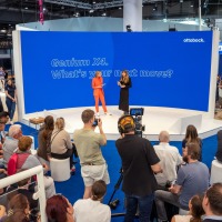 A stage with two speakers in front of a blue back wall, with a large audience and a cameraman in front of it.