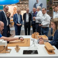 Two individuals demonstrating rehabilitation and therapy devices at a table, surrounded by a group of interested trade fair attendees in business attire.