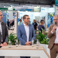 Three men in business attire, one speaking into a microphone, with exhibition booths and trade fair activity in the background.