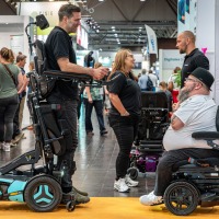 Two men in advanced electric wheelchairs conversing at OT-World, surrounded by other visitors and assistive device booths.