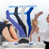 An exhibitor presents different colored compression stockings on side models. In the background, a woman reads a magazine.