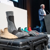 Innovative orthopedic foot and lower leg orthoses in various designs on a transport case, with a presentation taking place in the background at OTWorld