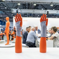 A table with four orange hand models showing different wrist bandages. Trade fair activity in the background.