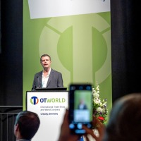 A speaker presents at the OTWorld Congress in Leipzig, while an attendee captures the scene with a smartphone.