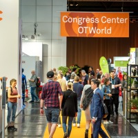 People walk through a trade fair hall with an orange carpet. In the background, a large orange banner reads “Congress Center OTWorld.” On the left side, there is a large advertising display featuring a woman. Attendees move between booths and engage in conversation.