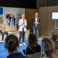 A woman speaks into a microphone on a small stage in front of a seated audience. A man stands next to her, while two other women are seated in the background. Medical compression stockings are displayed on mannequin legs mounted on the wall. A screen and a table with exhibit items are visible on the right.