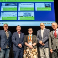 Five people stand side by side on a stage, facing the camera. A large screen behind them displays presentation slides related to orthopedic care. The individuals are wearing conference badges.