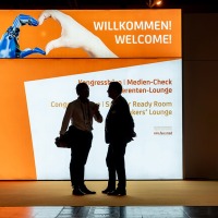 Two people are standing in silhouette in front of a large illuminated welcome screen that reads “WILLKOMMEN! WELCOME!” along with further directions to the congress area. At the top, an illustration shows a human and a bionic hand forming a heart shape.