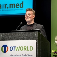 A speaker stands at a podium with the OTWorld logo, speaking at the international trade show. In the background, the logo of the company "con.fair.med Gesellschaft für Congressmanagement mbH" is visible.