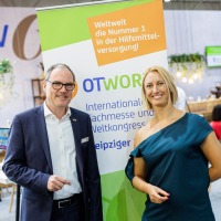 A man in a suit and a woman in a green dress are standing at OTWorld in front of a roll-up banner reading “OTWorld – International Trade Show and World Congress” and the slogan “Number 1 worldwide in assistive technology supply!”; in the background are other trade fair visitors and a lounge area.