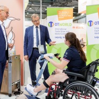 A young woman in a wheelchair uses a pedal-based therapy device at OTWorld, while two men in suits observe the equipment; in the background are two green OTWorld roll-up banners with English and German text.