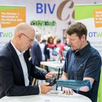 A man in a dark suit examines a blue-black arm prosthesis worn by another man in a dark shirt at OTWorld; in the background are two green OTWorld roll-up banners with English and German text.